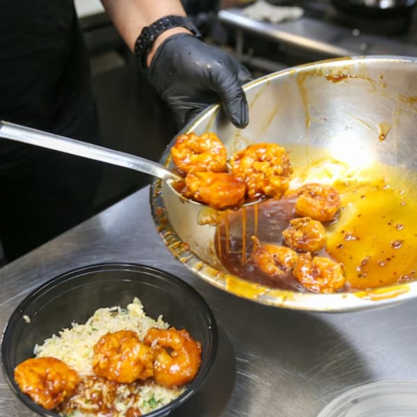 A chef in black gloves spoons orange glazed shrimp from a metal bowl onto a bed of rice.
