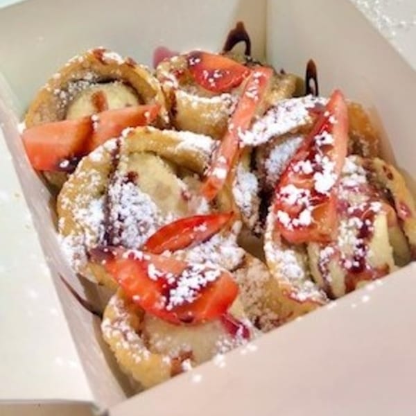 Fried banana fritters with powdered sugar, strawberry slices, and chocolate drizzle in a white box.