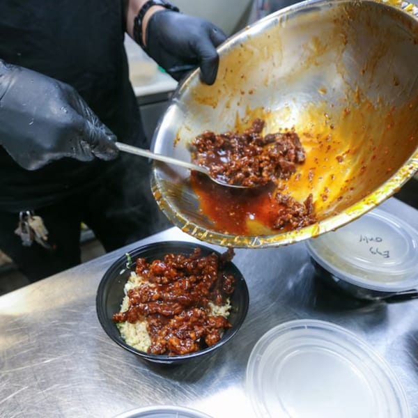 A chef in black gloves spoons saucy orange chicken from a metal bowl onto rice in a black takeout container.