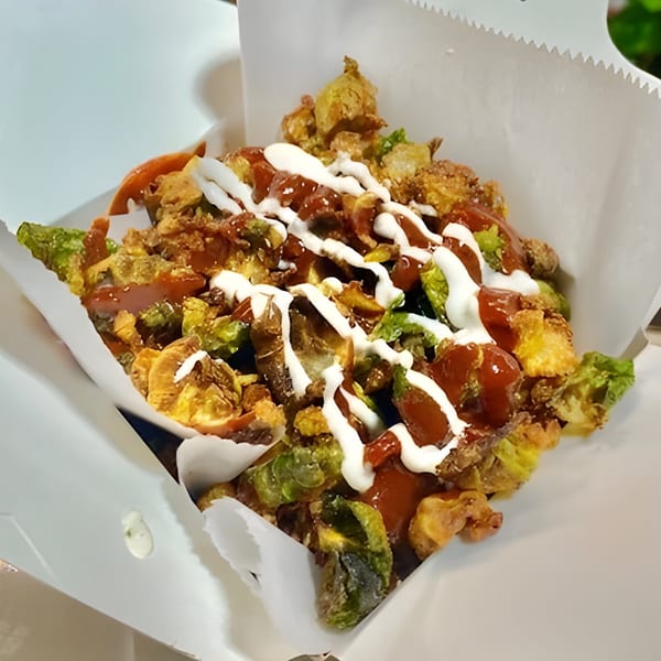 Crispy fried Brussels sprouts and other vegetables drizzled with red and white sauces in a white paper container.